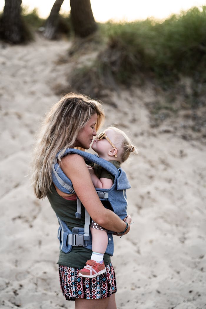 A loving mother kisses her toddler in a baby carrier on a sandy beach, capturing a tender parenting moment.