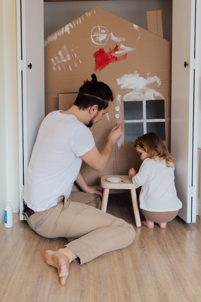Father and daughter painting a cardboard house indoors, enjoying quality time together.