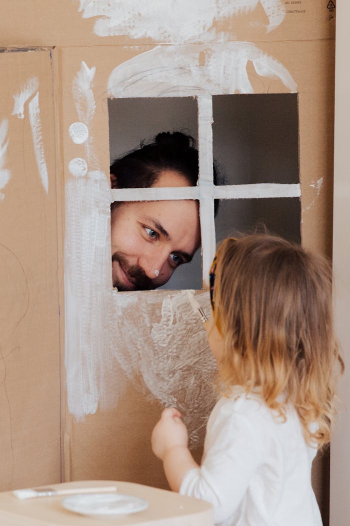 Father and child enjoy creative play with a cardboard house indoors.