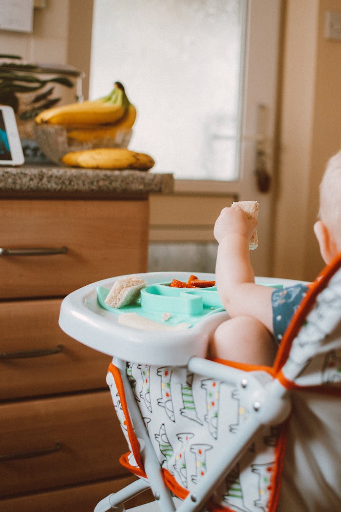 Baby in high chair eating food, surrounded by bananas in a kitchen setting.